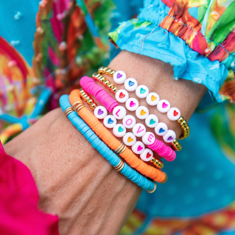 A wrist with five bracelets in the colors, pink, orange, and blue, against a floral backdrop. 