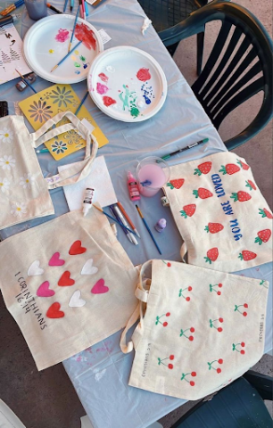 A table with a white tablecloth. On top of it, are three canvas tote bags each painted with their own designs. 